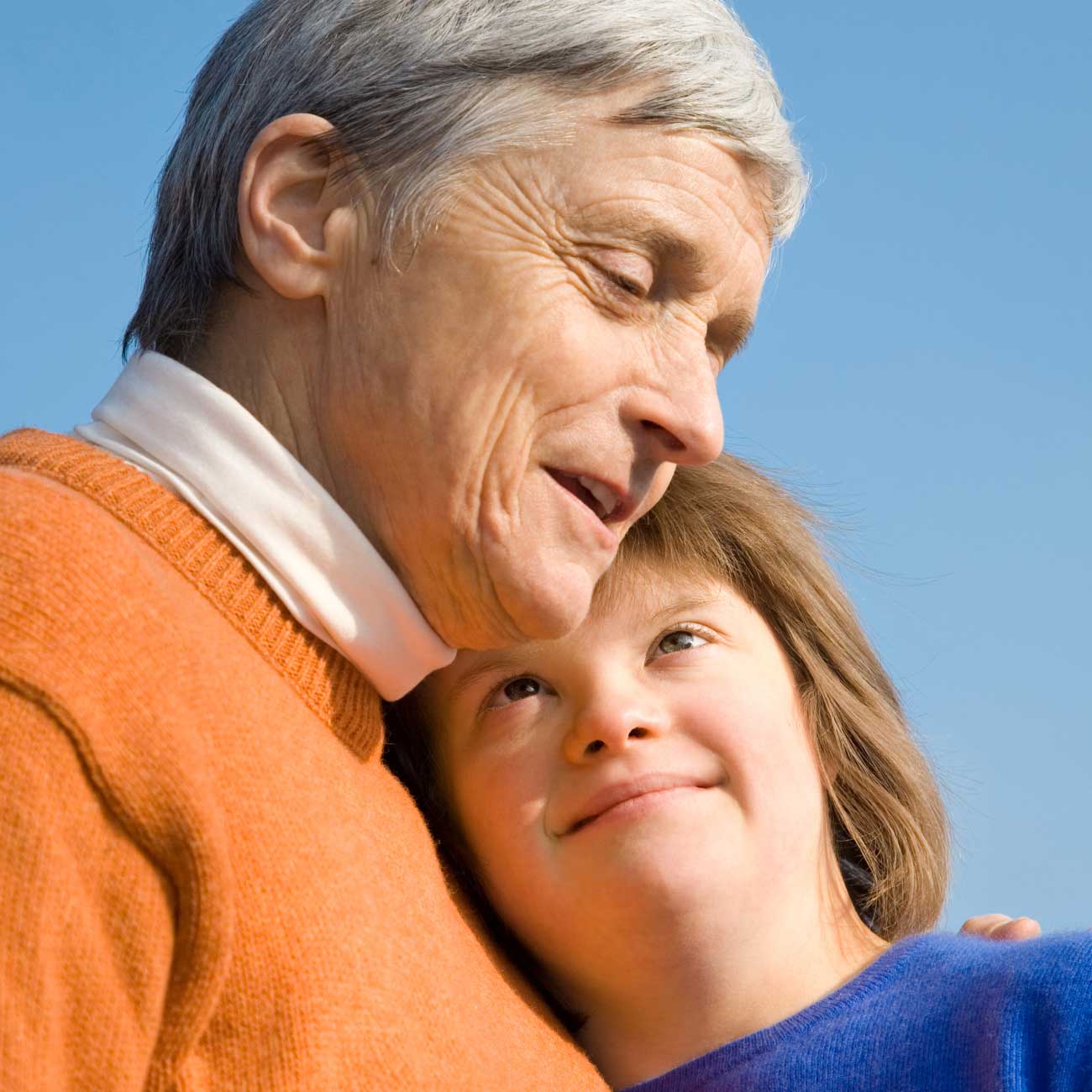 Grandparent embracing a young girl with Down syndrome while smiling under a clear blue sky