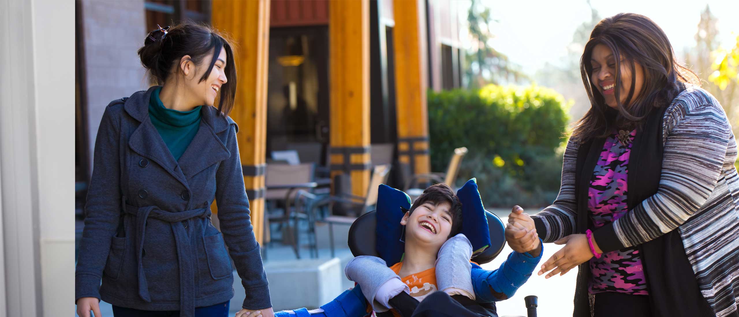 Two caregivers smiling and supporting a laughing child in a wheelchair outside a community care facility